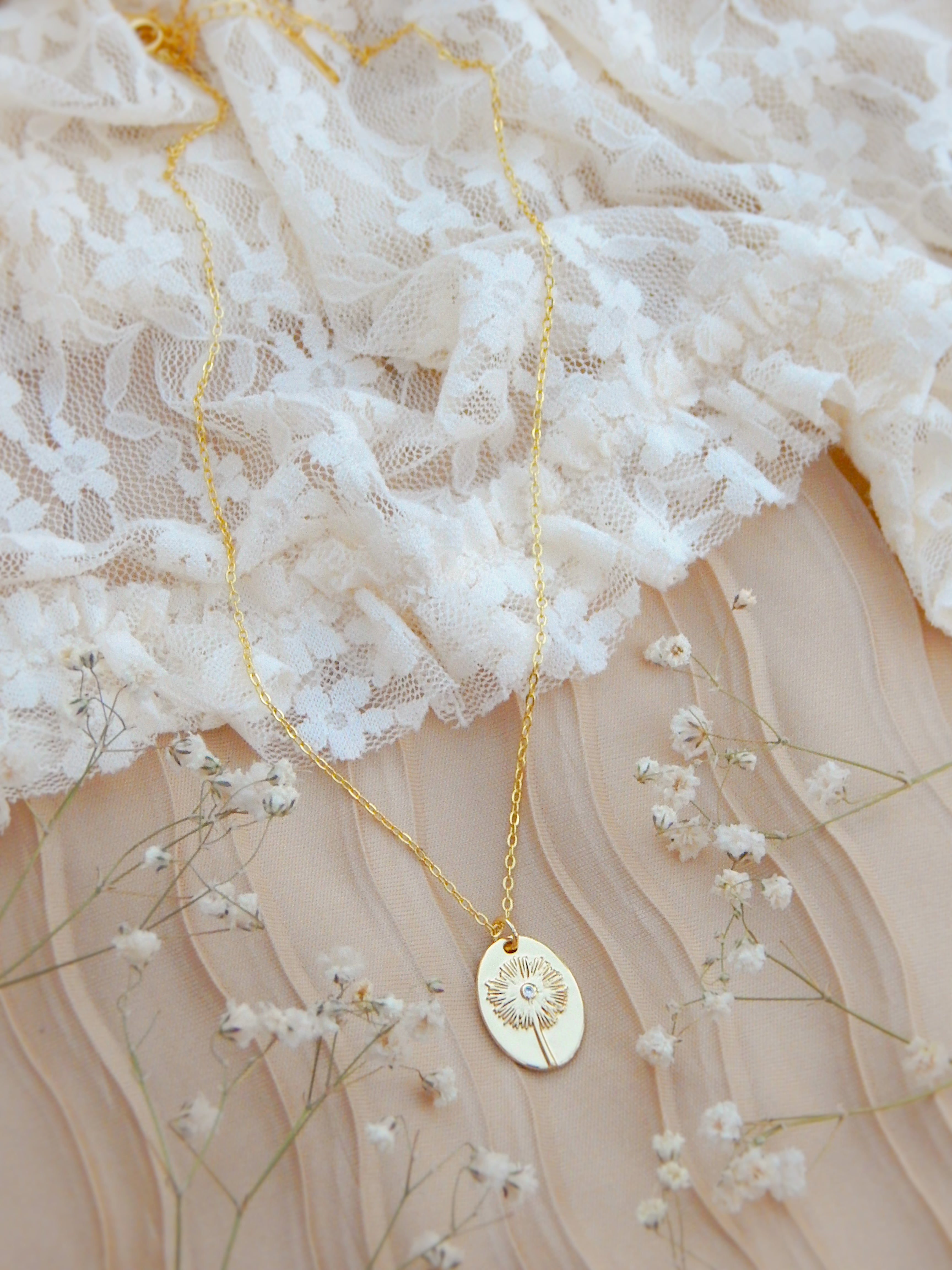 Gold dandelion pendant necklace styled on soft lace with delicate floral background