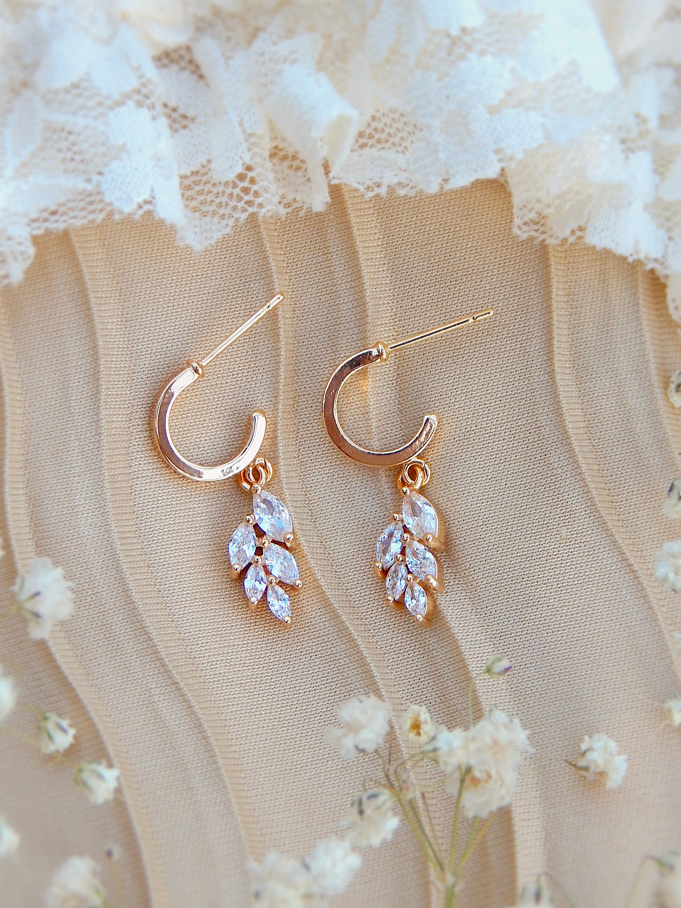 Gold huggie earrings with vine-inspired cubic zirconia leaf drop displayed on soft neutral fabric with floral accents