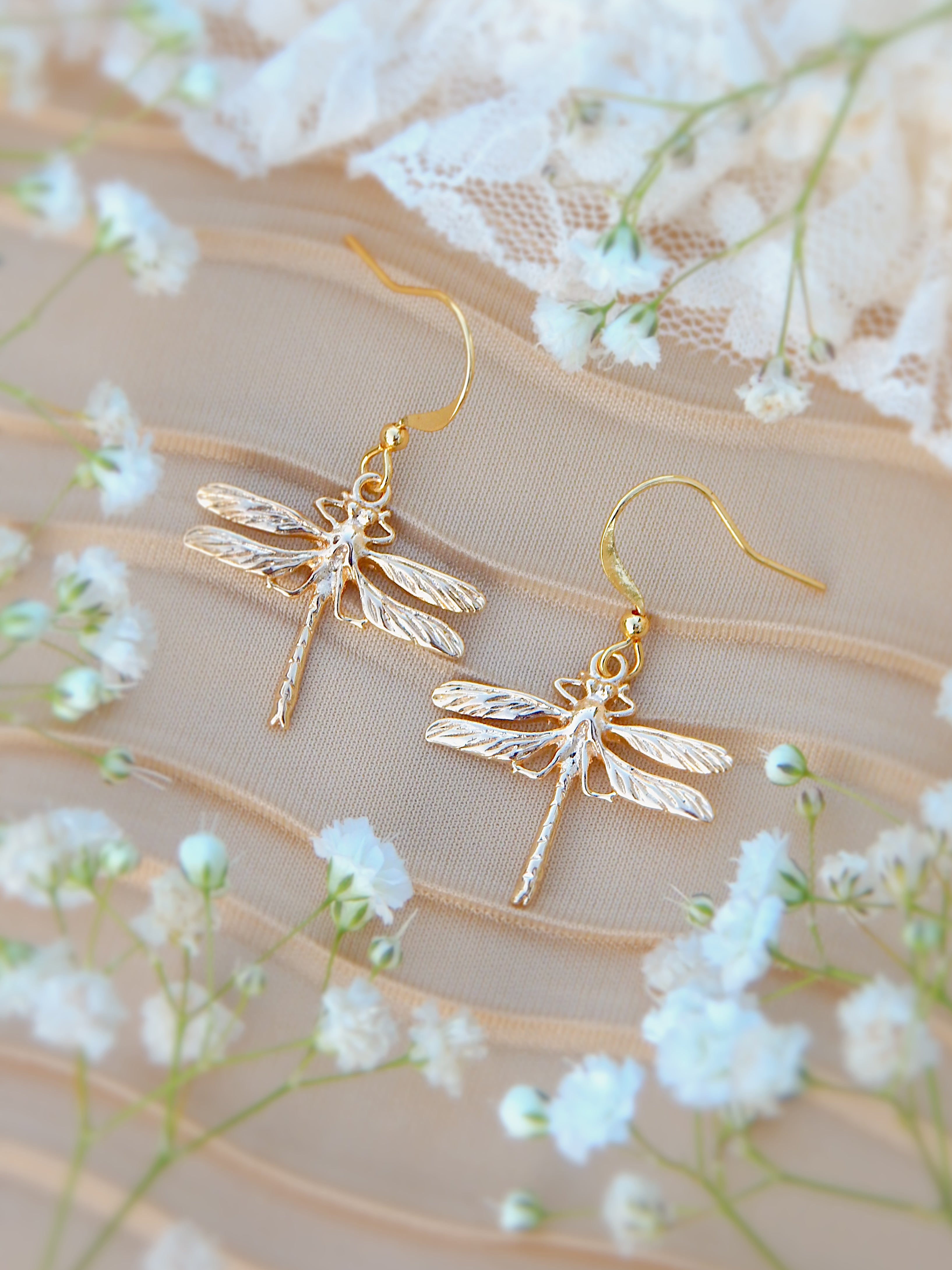 Gold dragonfly dangle earrings on neutral surface with soft white flowers, nature inspired jewelry
