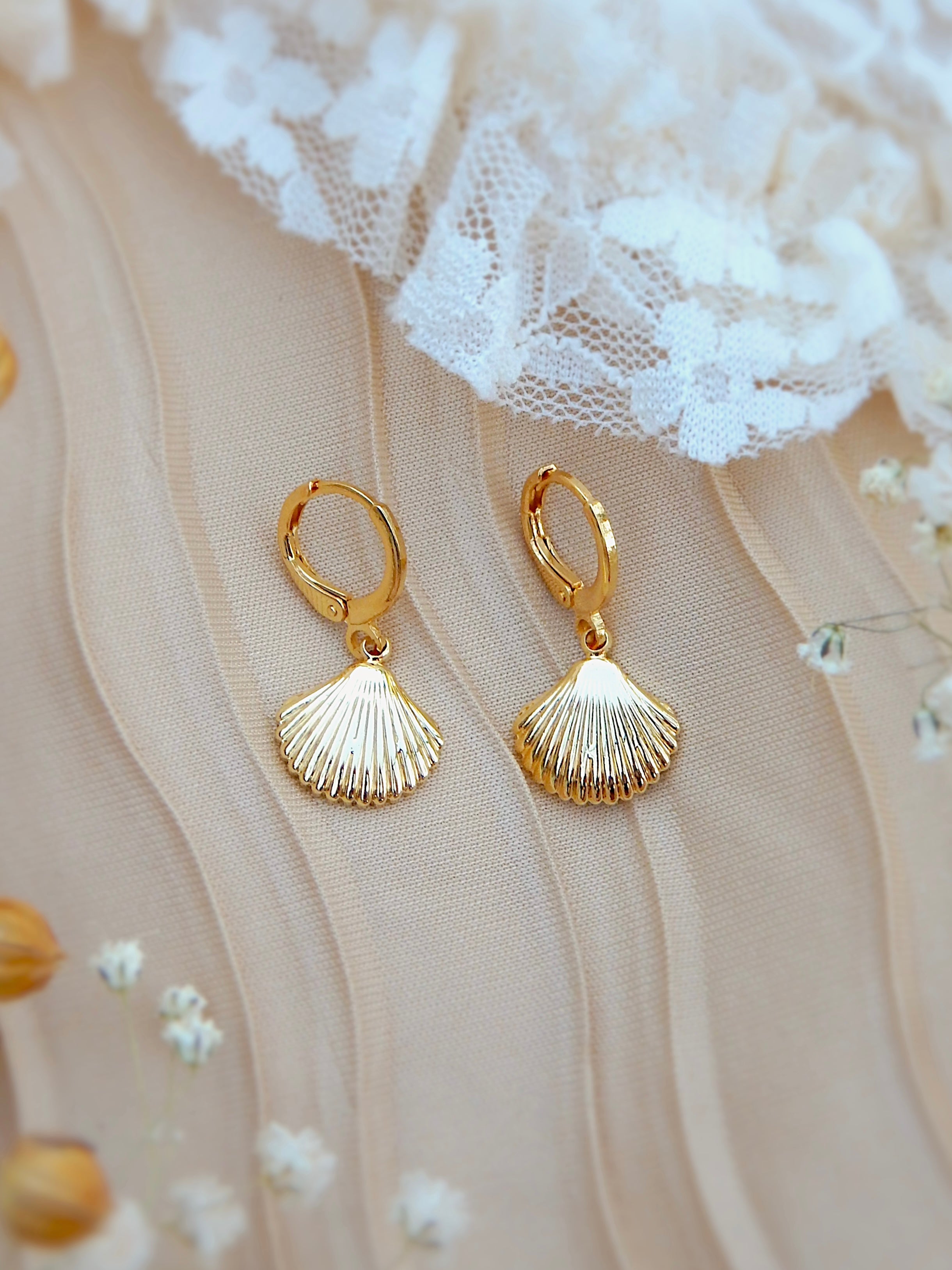 Gold seashell huggie earrings with textured shell charm displayed on soft neutral fabric with delicate floral accents
