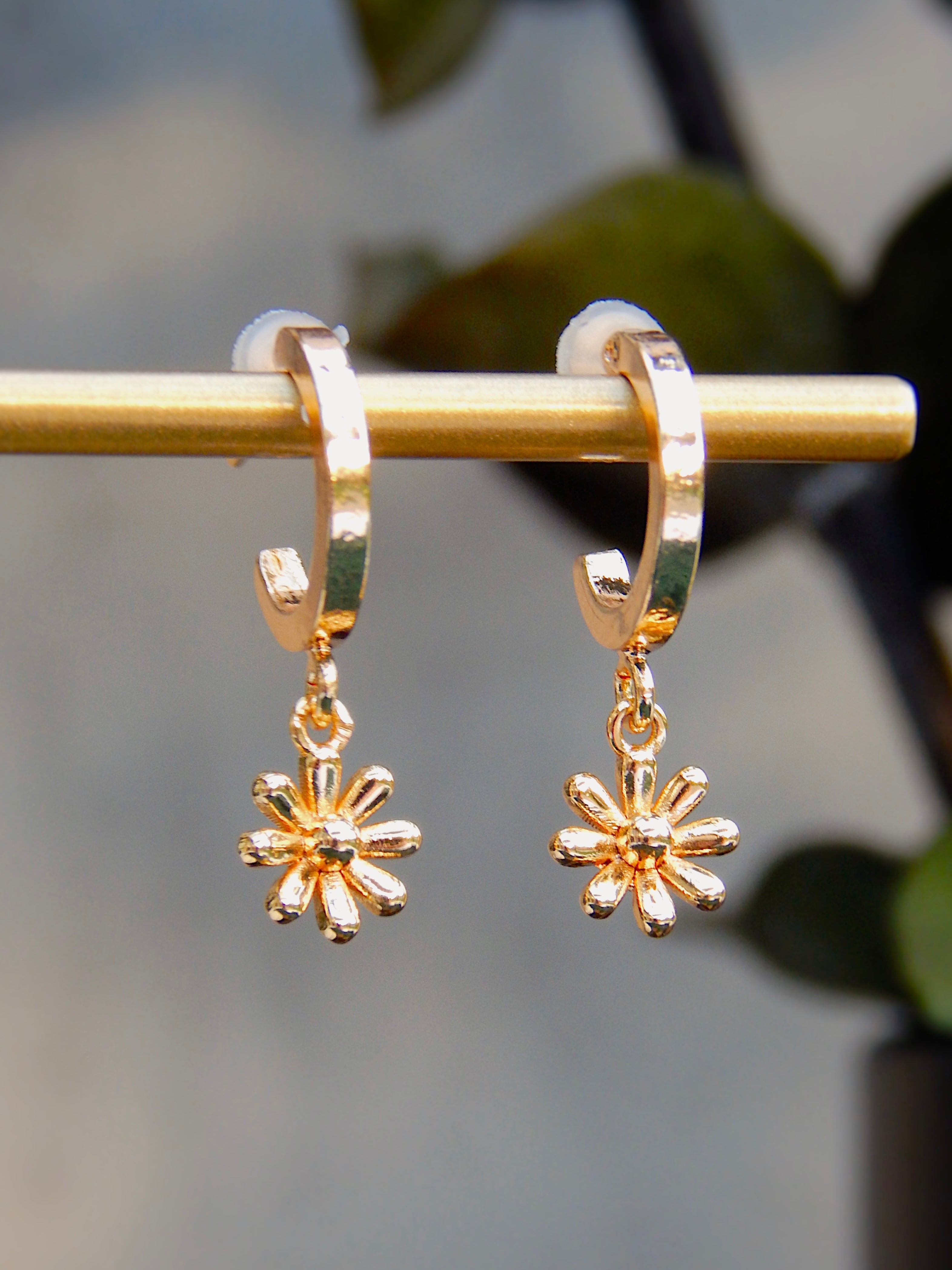 Gold floral huggie earrings with dainty flower charm hanging from small hoop, shown in close-up on jewelry stand