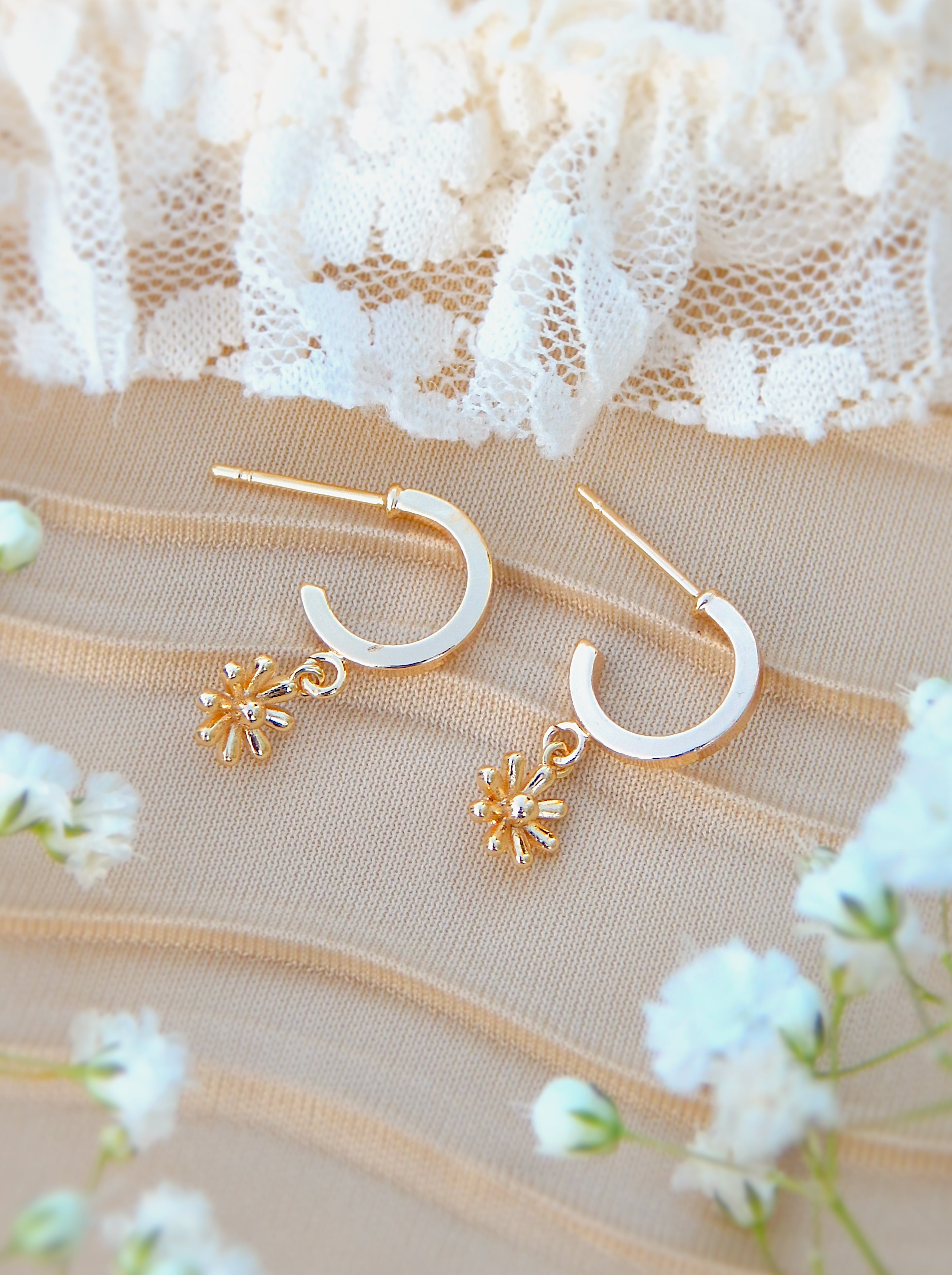 Gold floral huggie earrings with dainty flower charm displayed on soft neutral fabric with delicate flowers
